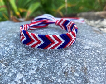 Puede incluir: Un brazalete de amistad rojo, blanco y azul con un patrón de chevron. El brazalete está hecho de cuerda tejida y tiene un cierre anudado.