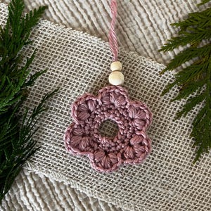 Puede incluir: Un adorno de flor de ganchillo en color rosa empolvado con una abertura circular central, seis pétalos redondeados y un lazo para colgar adornado con dos pequeñas cuentas de madera de color natural en hilo suave.