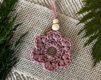 Puede incluir: Un adorno de flor de ganchillo en color rosa empolvado con una abertura circular central, seis pétalos redondeados y un lazo para colgar adornado con dos pequeñas cuentas de madera de color natural en hilo suave.