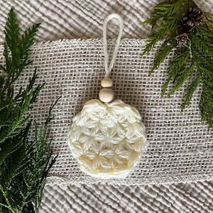 Puede incluir: Un adorno de ganchillo de color crema, de forma redonda, con un patrón texturizado en forma de estrella. El adorno está rematado con dos pequeñas cuentas de madera natural lisa y un lazo de hilo color crema para colgar, creando un diseño simple.