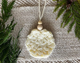 Puede incluir: Un adorno de ganchillo de color crema, de forma redonda, con un patrón texturizado en forma de estrella. El adorno está rematado con dos pequeñas cuentas de madera natural lisa y un lazo de hilo color crema para colgar, creando un diseño simple.