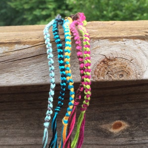 May include: Six colorful friendship bracelets made with braided string in shades of blue, green, pink, yellow, black, and gray.