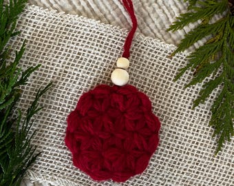 Puede incluir: Un adorno circular de ganchillo rojo con un patrón en forma de estrella, rematado con dos pequeñas cuentas de madera natural y un lazo de hilo rojo para colgar. La pieza está hecha de hilo grueso y muestra una textura cosida a mano.