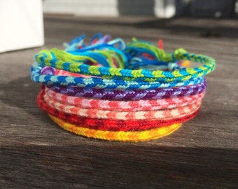 Puede incluir: Una pila de pulseras de amistad de colores en tonos de verde, azul, morado, rosa, rojo y amarillo. Las pulseras están hechas de cuerda tejida y están dispuestas en un patrón de arcoíris.