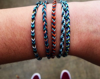 Puede incluir: Cinco pulseras de amistad trenzadas de colores en la muñeca de una persona. Las pulseras están hechas de hilos de diferentes colores y tienen un patrón de chevron.