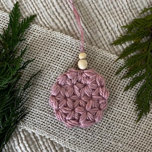 Puede incluir: Un adorno de ganchillo circular de color rosa polvoriento con un patrón texturizado en forma de estrella. El adorno está unido a un lazo corto de hilo rosa a juego y está decorado con dos pequeñas cuentas de madera de color natural en la parte superior.