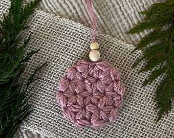 Puede incluir: Un adorno de ganchillo circular de color rosa polvoriento con un patrón texturizado en forma de estrella. El adorno está unido a un lazo corto de hilo rosa a juego y está decorado con dos pequeñas cuentas de madera de color natural en la parte superior.
