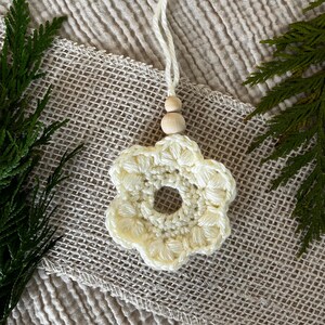 Puede incluir: Un adorno de flor de ganchillo color crema con una abertura circular central y seis pétalos redondeados. El adorno está sujeto a un lazo de cuerda blanca con dos pequeñas cuentas de madera redondas y claras situadas sobre la flor.
