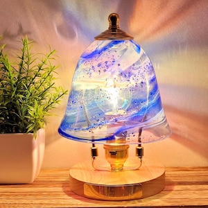 Puede incluir: Una pantalla de lámpara de vidrio azul con remolinos y burbujas blancas y azules se asienta sobre una base de madera con una placa de latón. La pantalla está iluminada con una bombilla blanca cálida.