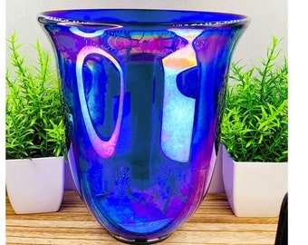 May include: A large, iridescent blue glass vase with a flared opening and a rounded bottom. The vase has a pearlescent finish that shimmers with shades of purple and pink.