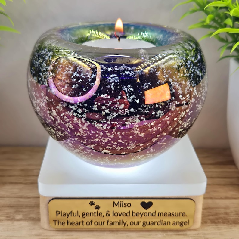 May include: A lit candle in a decorative glass orb with a multi-colored, iridescent finish. The orb sits on a white base with a wooden bottom. Text on the base reads: "Miiso, Playful, gentle, & loved beyond measure. The heart of our family, our guardian angel."