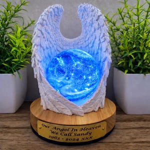 Puede incluir: Un adorno conmemorativo con alas de ángel esculpidas en blanco que sostienen una esfera azul brillante con luces internas. El objeto se asienta sobre una base circular de madera con una placa dorada grabada con el texto: Our Angel In Heaven We Call Sandy 1963 - 2024 XXX.