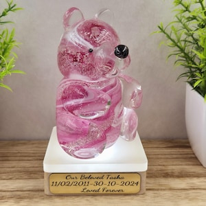 Puede incluir: Figurita de oso de vidrio rosa con remolinos blancos y ojos negros, sentada sobre una base blanca con una placa de madera que dice "Our Beloved Tasha 11/02/2011-30-10-2024 Loved Forever".