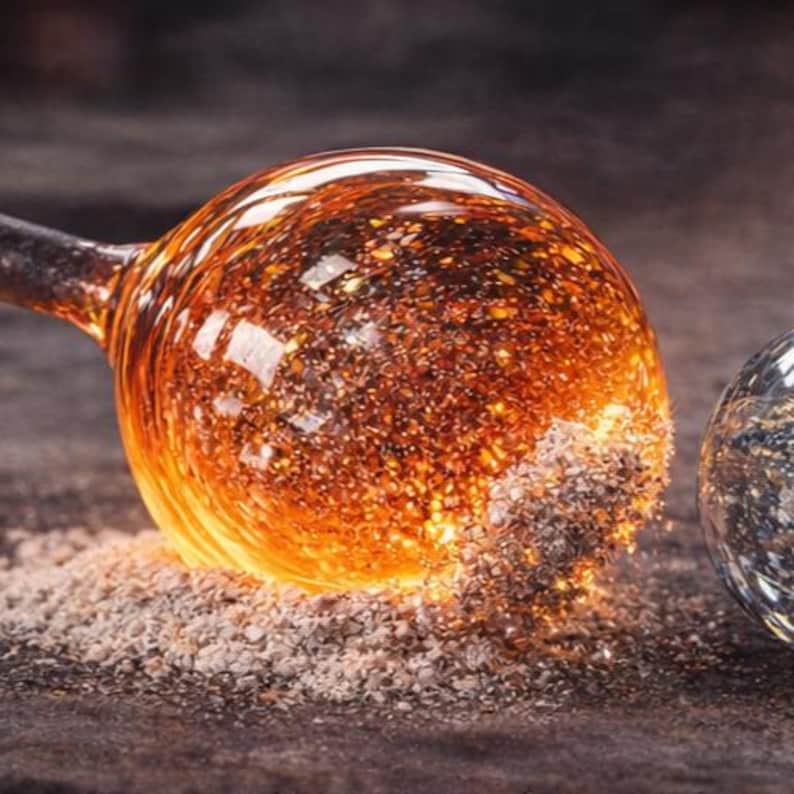May include: Close-up of a glass art piece with a round, amber-colored orb. The orb has a textured surface and is partially covered in a fine, gray powder. A second, clear glass orb is visible in the background.