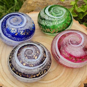May include: Four glass spheres in shades of blue, green, pink, and black, featuring a swirling white and gold glitter pattern. The decorative spheres are presented on a wooden surface, highlighting their vibrant colours and textures.