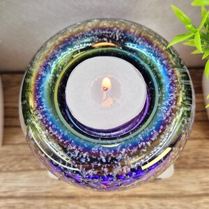 May include: A round, iridescent glass candle holder with a lit white tea light candle inside. The glass has a speckled, textured appearance with colors of blue, purple, and green. The candle holder sits on a white base.
