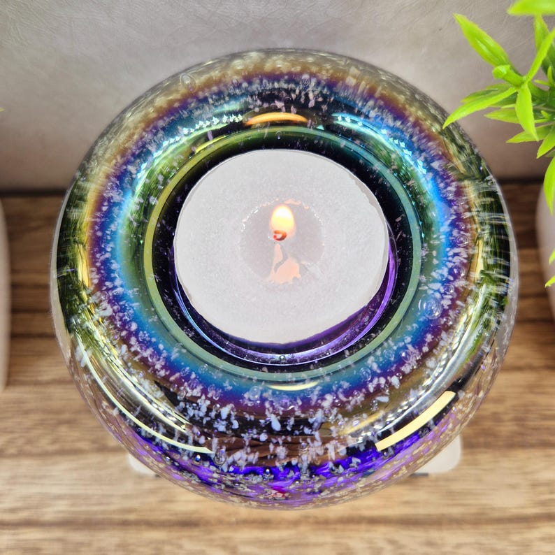 May include: A round, iridescent glass candle holder with a lit white tea light candle inside. The glass has a speckled, textured appearance with colors of blue, purple, and green. The candle holder sits on a white base.