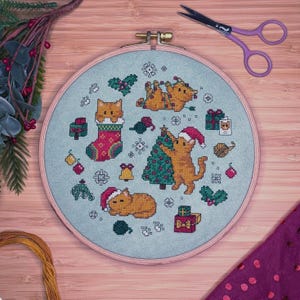 May include: A round wooden embroidery hoop containing light blue fabric with a cross-stitch pattern of orange cats, Christmas trees, presents, and snowflakes. A pair of purple scissors is positioned in the upper right corner of the frame.