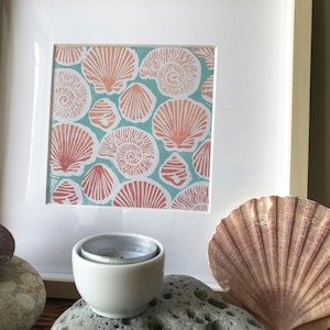 May include: A framed print of a seashell pattern in shades of pink and orange on a teal background. The print is surrounded by a white mount and a light wood frame.