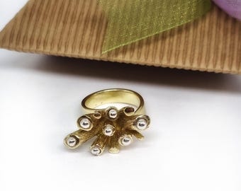 May include: A gold-colored ring with a unique design. The ring features a cluster of small, silver-colored spheres attached to gold-colored, branch-like structures. The ring is set against a white background, with a gift box and ribbon in the background.