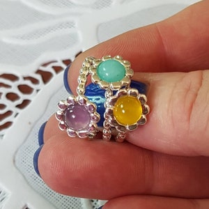 Può includere: Tre anelli d'argento con pietre colorate. Gli anelli sono impilati uno sopra l'altro. Le pietre sono viola, verdi e gialle.