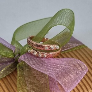 May include: A hammered copper ring with a silver band and silver beads. The ring is wrapped around a green and purple ribbon.