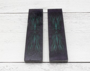 May include: Two rectangular, dark purple pieces with a teal tribal design. The design is symmetrical and centered on each piece. The background is a white, wooden surface.