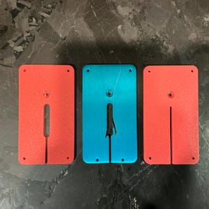 May include: Three rectangular metal plates: two red and one blue. Each plate has a central slot and a circular hole. The plates are likely components of a larger device or system.