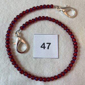 Könnte beinhalten: Eine rote und blaue Perlenkette mit silbernen Verschlüssen. Die Kette hat ein Etikett mit der Nummer 47.