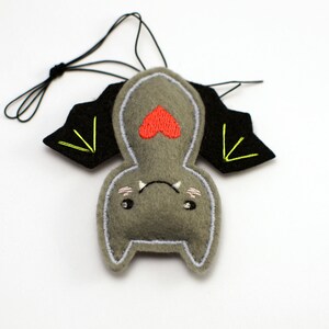 Op de afbeelding: Een grijs vilten vleermuisornament met zwarte vleugels en een rood hart. De vleermuis heeft een witte omtrek, geborduurde details en een zwart touwtje om op te hangen. De vleugels hebben gele stiksels.