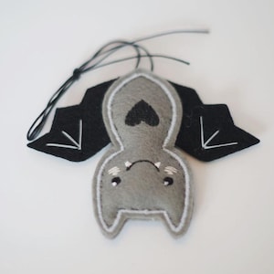 Op de afbeelding: Een grijs vilten vleermuisornament met zwarte vleugels en een zwart touwtje om op te hangen. De vleermuis heeft witte geborduurde details, waaronder een hart op de borst en accenten op de vleugels. Het gezicht van de vleermuis heeft geborduurde ogen en een kleine mond.
