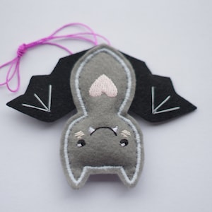 Op de afbeelding: Een grijs vilten vleermuisornament met zwarte vleugels en een roze hartvormige neus. De vleermuis heeft witte geborduurde details en een paarse draad om op te hangen. Een schattige Halloween-decoratie.