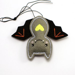 Op de afbeelding: Een grijs vilten vleermuisornament met zwarte vleugels en oranje stiksels. De vleermuis heeft een gele hart op de borst en een klein, lachend gezicht. Een zwart touwtje is bevestigd om op te hangen.