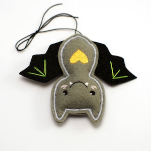 Op de afbeelding: Een grijs vilten vleermuisornament met zwarte vleugels en een gele hart op de borst. De vleermuis heeft een witte omtrek, geborduurde details en een zwart touwtje om op te hangen. De vleugels hebben groene geborduurde accenten.