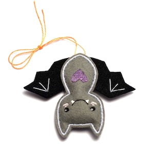 Op de afbeelding: Een grijs vilten vleermuisornament met zwarte vleugels en witte stiksels. De vleermuis heeft een paars hart en een oranje touwtje. Een Halloween decoratie.