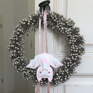 Op de afbeelding: Een zilveren bessenkrans met een hangend wit vilten vleermuisornament. De vleermuis heeft roze accenten en rode geborduurde details. Een roze lint wordt gebruikt om het ornament en de krans aan een witte deur op te hangen.
