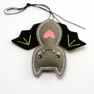 Op de afbeelding: Een grijs vilten vleermuisornament met zwarte vleugels en neongele stiksels. De vleermuis heeft een roze hart op de borst en geborduurde details. Een zwart touwtje is bevestigd om op te hangen.
