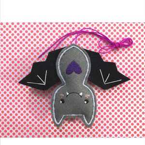 Op de afbeelding: Een grijs vilten vleermuisornament met zwarte vleugels en een paars hartdetail. De vleermuis heeft witte stiksels en een paars touwtje om op te hangen. De achtergrond is een rood-wit stippenpatroon.