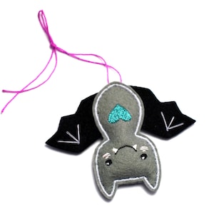 Op de afbeelding: Een grijze vilten vleermuis met zwarte vleugels en een turquoise hartdetail. De vleermuis heeft witte stiksels en een roze touwtje om op te hangen. Een schattige, handgemaakte Halloween-decoratie.