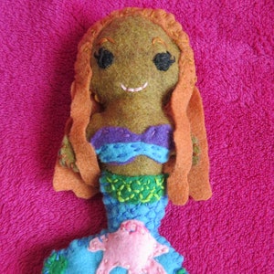 May include: A felt mermaid doll with long orange hair, a purple and blue top, a green and blue tail, and a pink starfish on the tail.