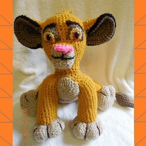 May include: A crocheted stuffed lion toy with brown ears, a yellow body, and a pink nose. The lion is sitting on a white fluffy surface.