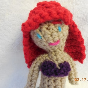 May include: Crocheted doll with red hair, blue eyes, and a purple top. The doll is made of yarn and has a simple, handmade look.