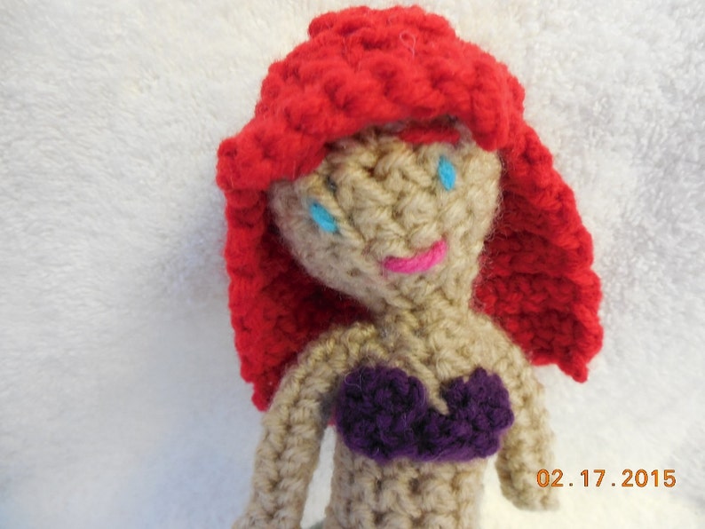May include: Crocheted doll with red hair, blue eyes, and a purple top. The doll is made of yarn and has a simple, handmade look.