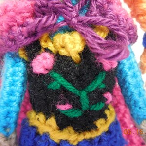 May include: Crocheted doll with a purple, pink, blue, and yellow outfit. The doll has a black vest with green vines and pink flowers. The doll's hair is orange.