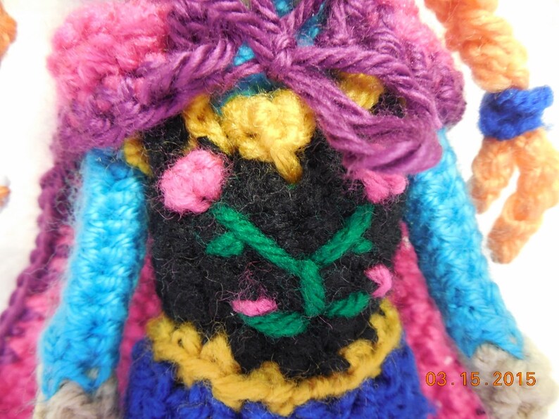May include: Crocheted doll with a purple, pink, blue, and yellow outfit. The doll has a black vest with green vines and pink flowers. The doll's hair is orange.