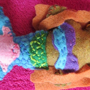 May include: A felt mermaid doll with a pink, blue, green, and purple bodice. The mermaid is wearing a brown felt cloak.