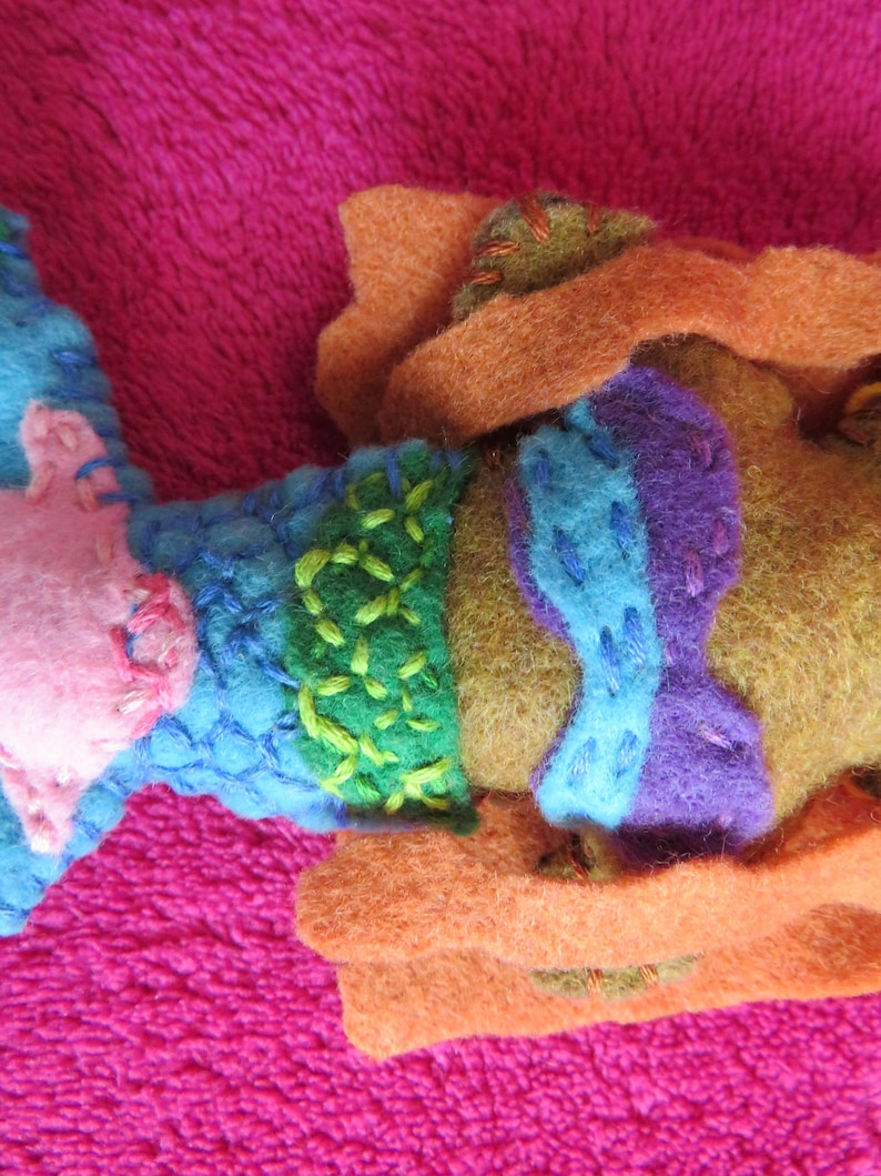 May include: A felt mermaid doll with a pink, blue, green, and purple bodice. The mermaid is wearing a brown felt cloak.