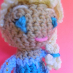 Op de afbeelding: Een gehaakte pop met blond haar, blauwe ogen en een roze mond. De pop draagt een blauwe jurk met witte accenten. De pop is gemaakt van garen en is ongeveer 25 cm hoog.