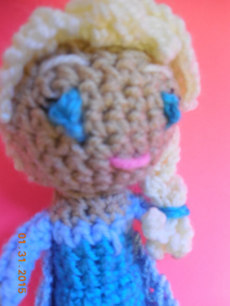 Op de afbeelding: Een gehaakte pop met blond haar, blauwe ogen en een roze mond. De pop draagt een blauwe jurk met witte accenten. De pop is gemaakt van garen en is ongeveer 25 cm hoog.