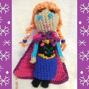 May include: Crocheted doll with red, purple, and blue clothing. The doll has orange hair in braids and blue eyes. The doll is wearing a cape and a dress with a floral design.